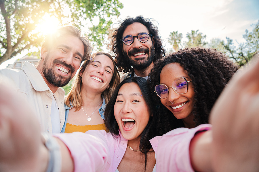 A multi-enthnic group of 5 young adults smiling as they take a group selfie together.